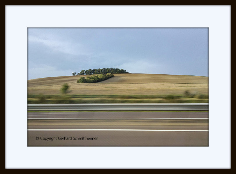 Landschaftsfoto aus dem fahrenden Auto mit linienartiger horizontaler Bewegungsunschärfe im Vordergrund und ruhiger Landschaft mit kleinem Wäldchen im Hintergrund, minimalistische Farbigkeit