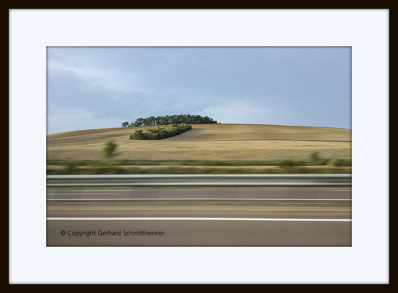 Landschaftsfoto aus dem fahrenden Auto  mit linienartiger horizontaler Bewegungsunschärfe im Vordergrund und ruhiger Landschaft mit kleinem Wäldchen im Hintergrund, minimalistische Farbigkeit
